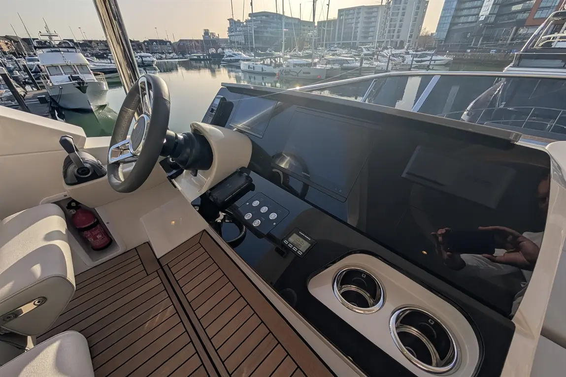 Boat cockpit with steering wheel, electronic displays, control panel, and wooden flooring, overlooking a marina at sunrise.